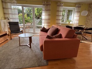 A living room with a sofa, a small table, a chair, and a dining table with chairs. There is a view of a garden through a glass door. - Apartment 'Findus' with Private Terrace and Wi-Fi (Kempten-Haldenwang)