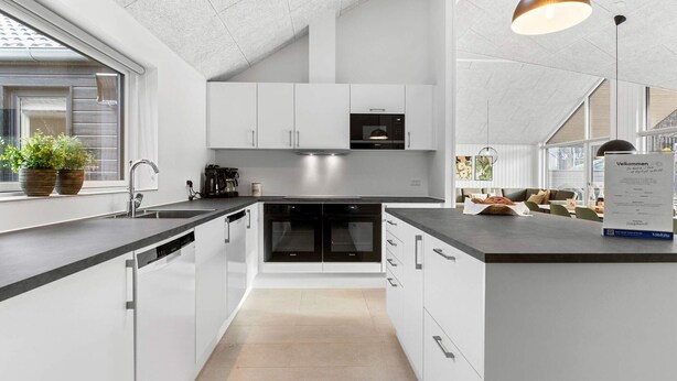 A modern kitchen with white cabinets, a black countertop, and a built-in oven. There are potted plants on the windowsill and a menu stand on the island. - 5 Star Holiday Home in Glesborg (Glesborg)