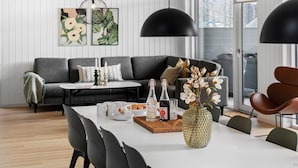 A modern living room with a dining area, featuring a sofa, a coffee table, and a dining table with chairs. There are decorative items on the table, including a vase with flowers and a tray with pastries. - 5 Star Holiday Home in Glesborg (Glesborg)