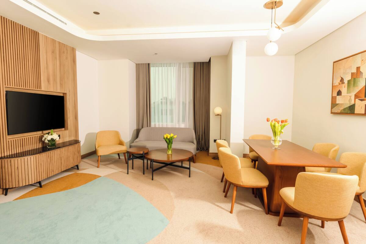 A modern hotel room with a flat-screen TV, a sofa, a dining table with chairs, and a large window with curtains.