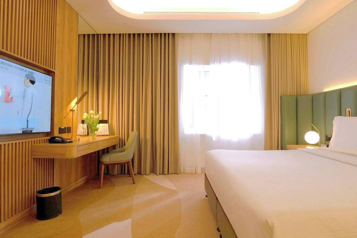 A hotel room with a bed, a desk with a computer, a chair, a TV, and a window with curtains.
