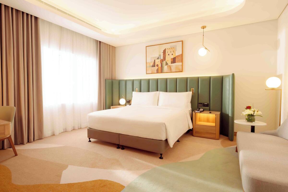 A hotel room with a large bed, a desk, a chair, a small table with a vase of flowers, and a painting on the wall.