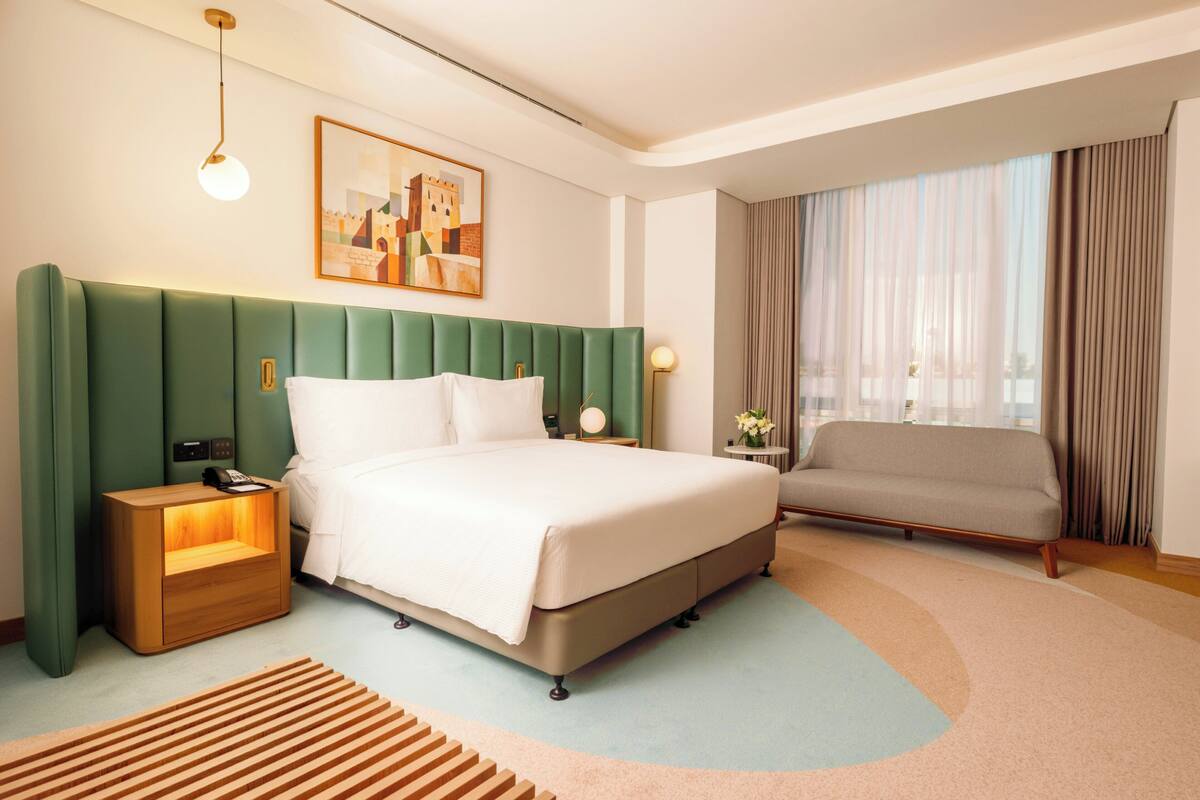 A hotel room with a green upholstered headboard, a bed with white linens, a bedside table with a lamp, a sofa, and a large window with curtains.