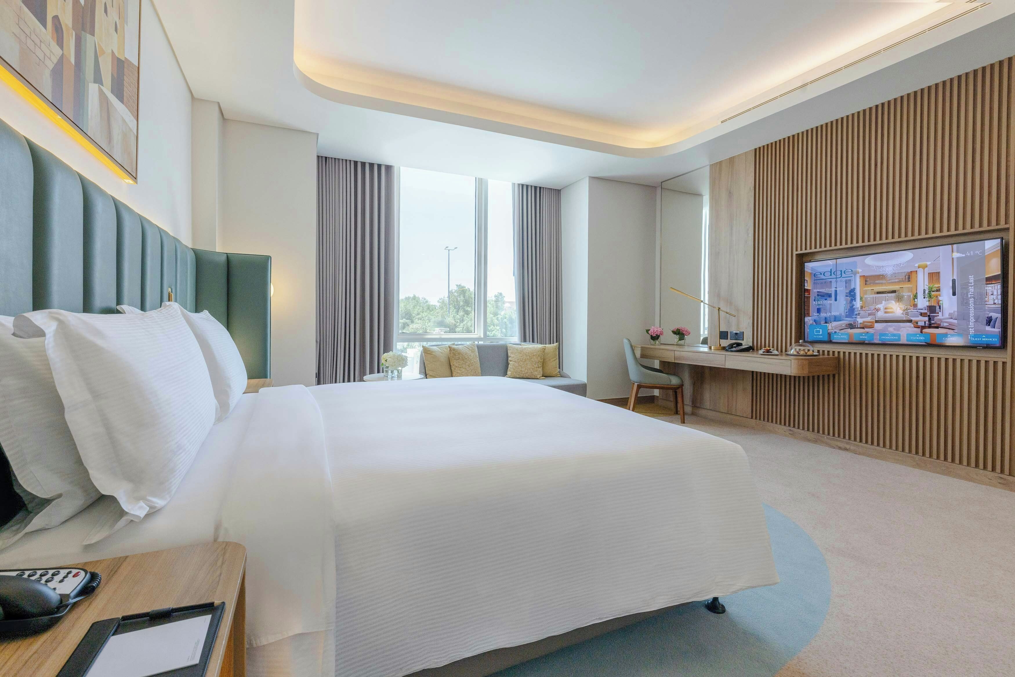 Habitación de hotel con una cama grande, un escritorio con computadora, televisión y ventana con cortinas.