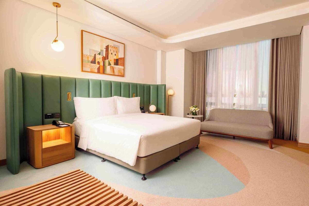 A hotel room with a green upholstered headboard, a bed with white bedding, a bedside table with a lamp, a sofa, and a large window with curtains.