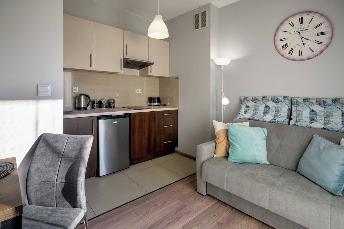 Una cocina moderna con lavavajillas, microondas y hervidor. Hay un sofá gris con cojines azules y beige, y un sillón gris. Se ve un reloj de pared grande.
