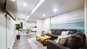 A modern living room with a dark brown sofa, a wooden coffee table, and a large window with a view of the ocean. - Sanctuary Suite - KingBed - Wifi - Near WEM/Rogers (Edmonton)