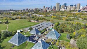 A solar farm with multiple triangular solar panels in a green field, surrounded by trees and a city skyline in the background. - Sanctuary Suite - KingBed - Wifi - Near WEM/Rogers (Edmonton)
