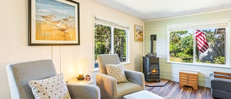 A cozy living room with two grey armchairs, a small ottoman, a fireplace, and a large window with an American flag.