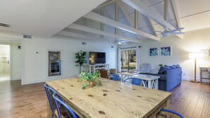 Amplia sala de estar con una mesa de comedor de madera, sillas azules, un ventilador de techo y un ventanal que proporciona luz natural.