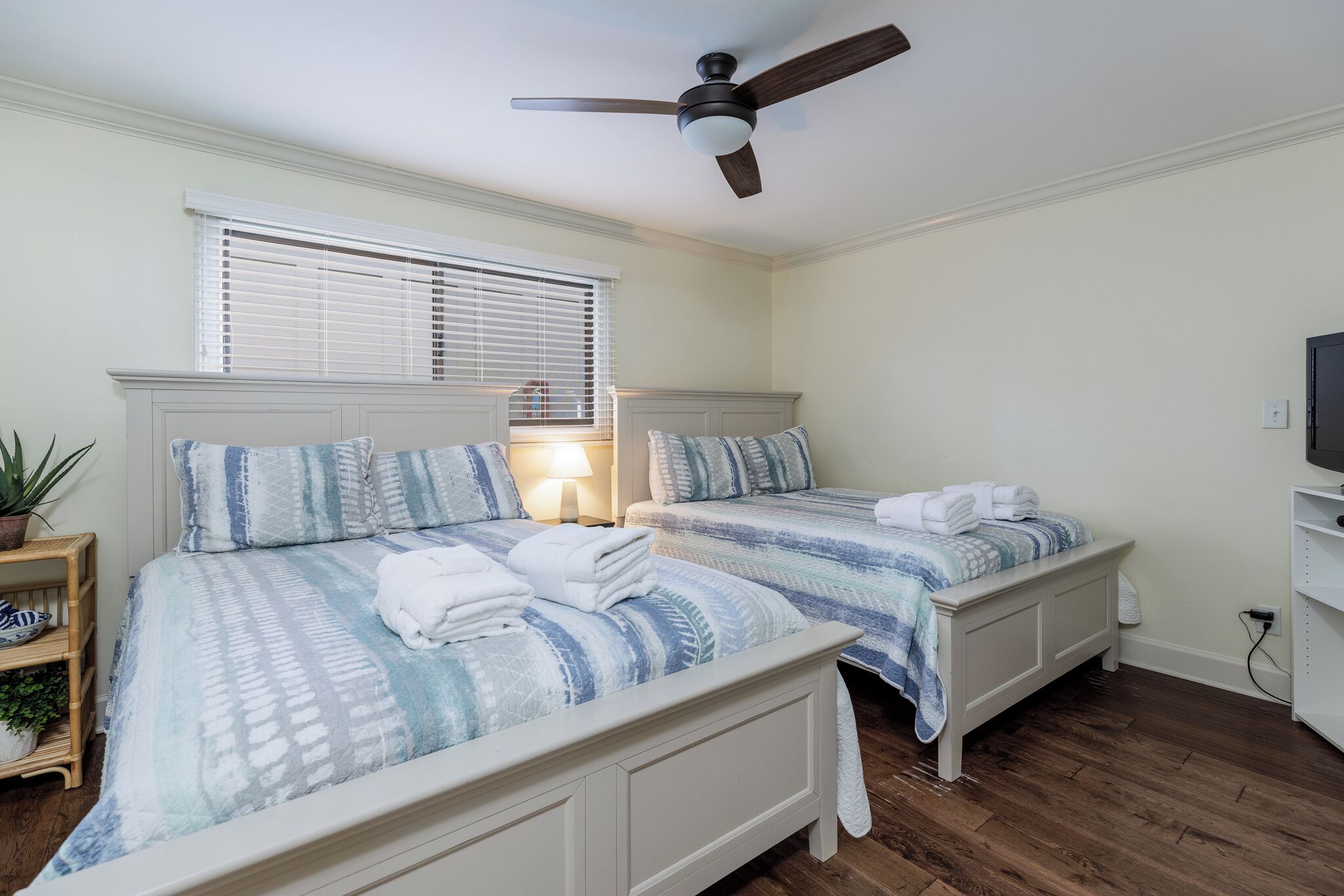 A bedroom with two beds, a ceiling fan, a window with blinds, a bedside lamp, and a TV on a shelf.