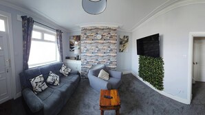 A living room with a sofa, armchair, and a small wooden table. There is a TV mounted on the wall and a decorative wall panel. - Charming 3-bedroom Home in Doncaster, Perfect for Family, Friends & Contractors (Doncaster)