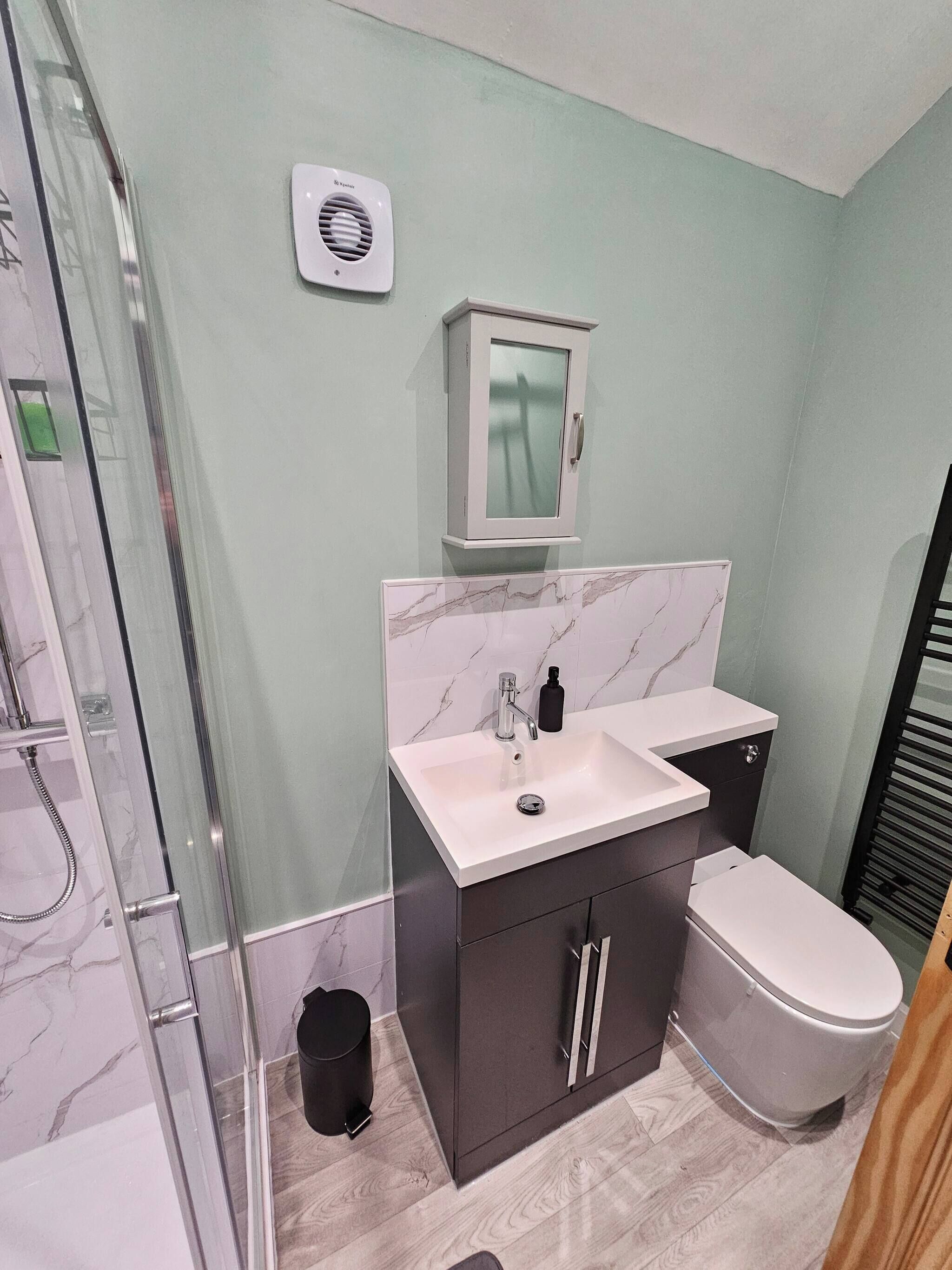 Een moderne badkamer met een douchecabine van glas, een witte wastafel met een marmeren achterwand, een toilet en een kast met een matglazen deur.