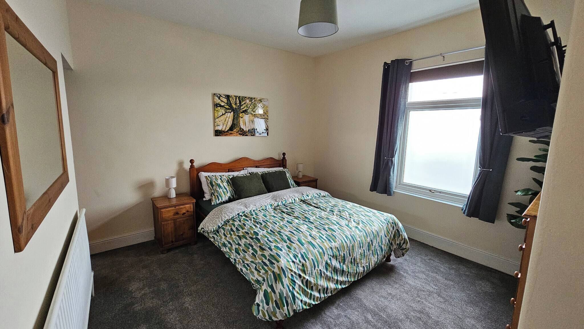 A bedroom with a bed, a nightstand, a wall art piece, a window with curtains, and a radiator.