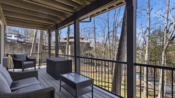 A covered outdoor deck with wicker furniture, a glass table, and a view of trees and a road. - Grand Ridge Retreat w Decks, TVs & Pool Access! (Osage Beach)