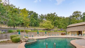 A swimming pool with lounge chairs and umbrellas, surrounded by trees and a building. - Grand Ridge Retreat w Decks, TVs & Pool Access! (Osage Beach)