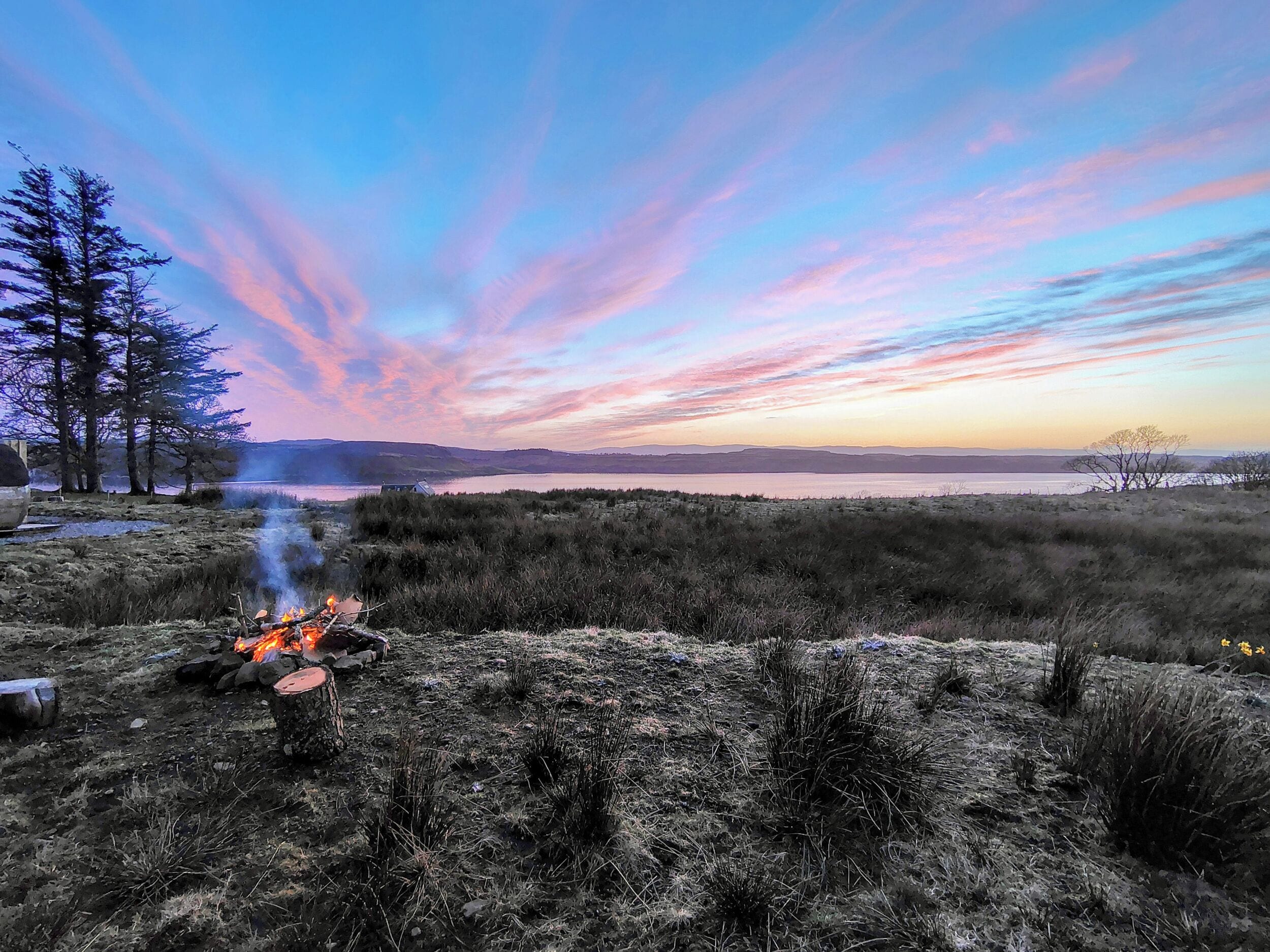 A campfire in a natural setting with trees and a distant view of a lake.