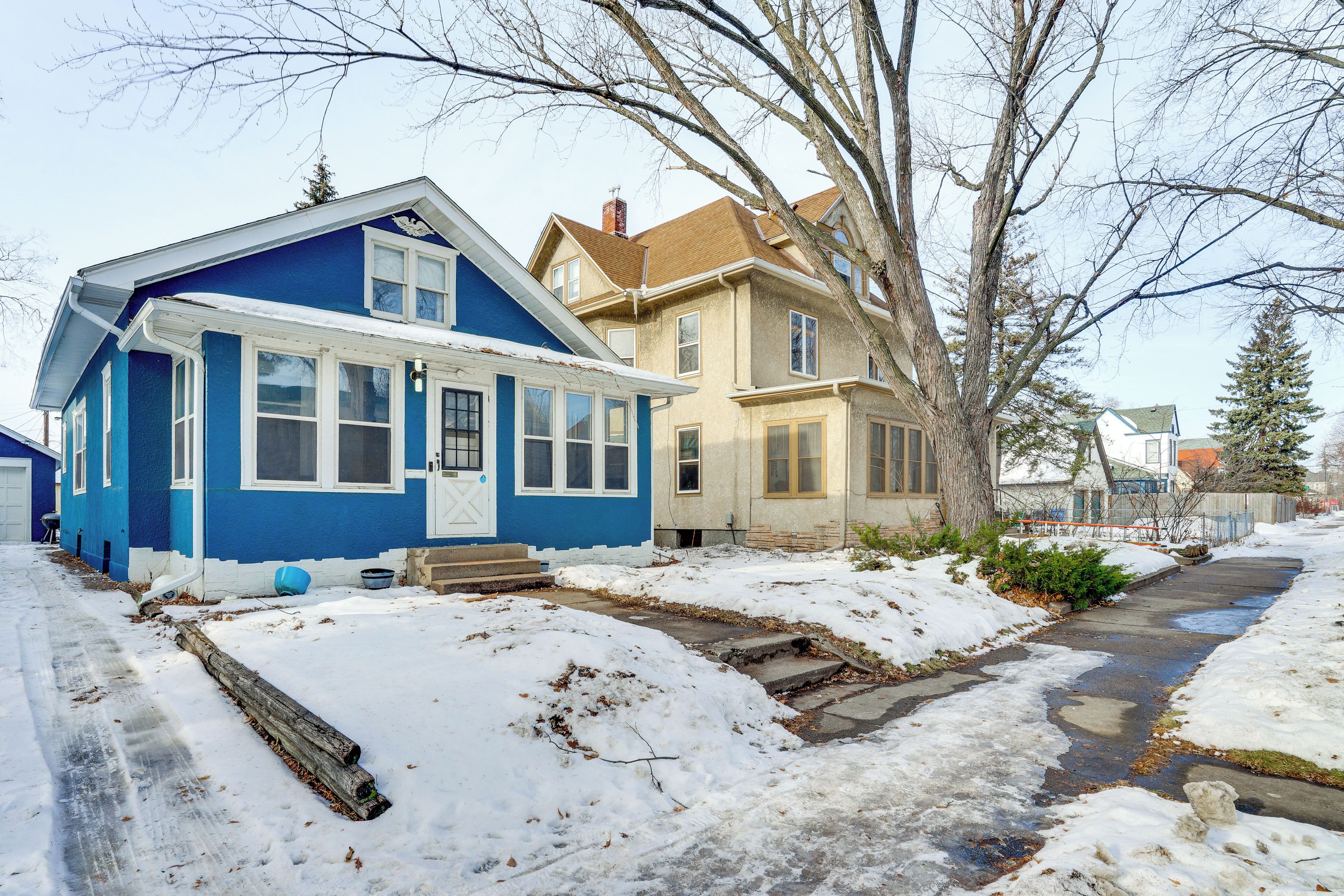 A residential street with two houses, one blue and one beige, with a snow-covered sidewalk and a small stream running through.