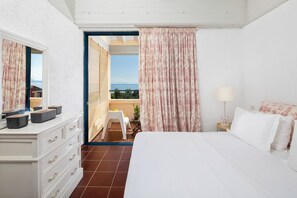 Ein Hotelzimmer mit Bett, Schminktisch, Sessel, kleinem Tisch mit Lampe und Meerblick durch eine Tür.