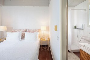 Une chambre d’hôtel comprenant un lit, des lampes de chevet, une petite table avec une lampe, un espace douche et une salle de bain avec un lavabo et un miroir.