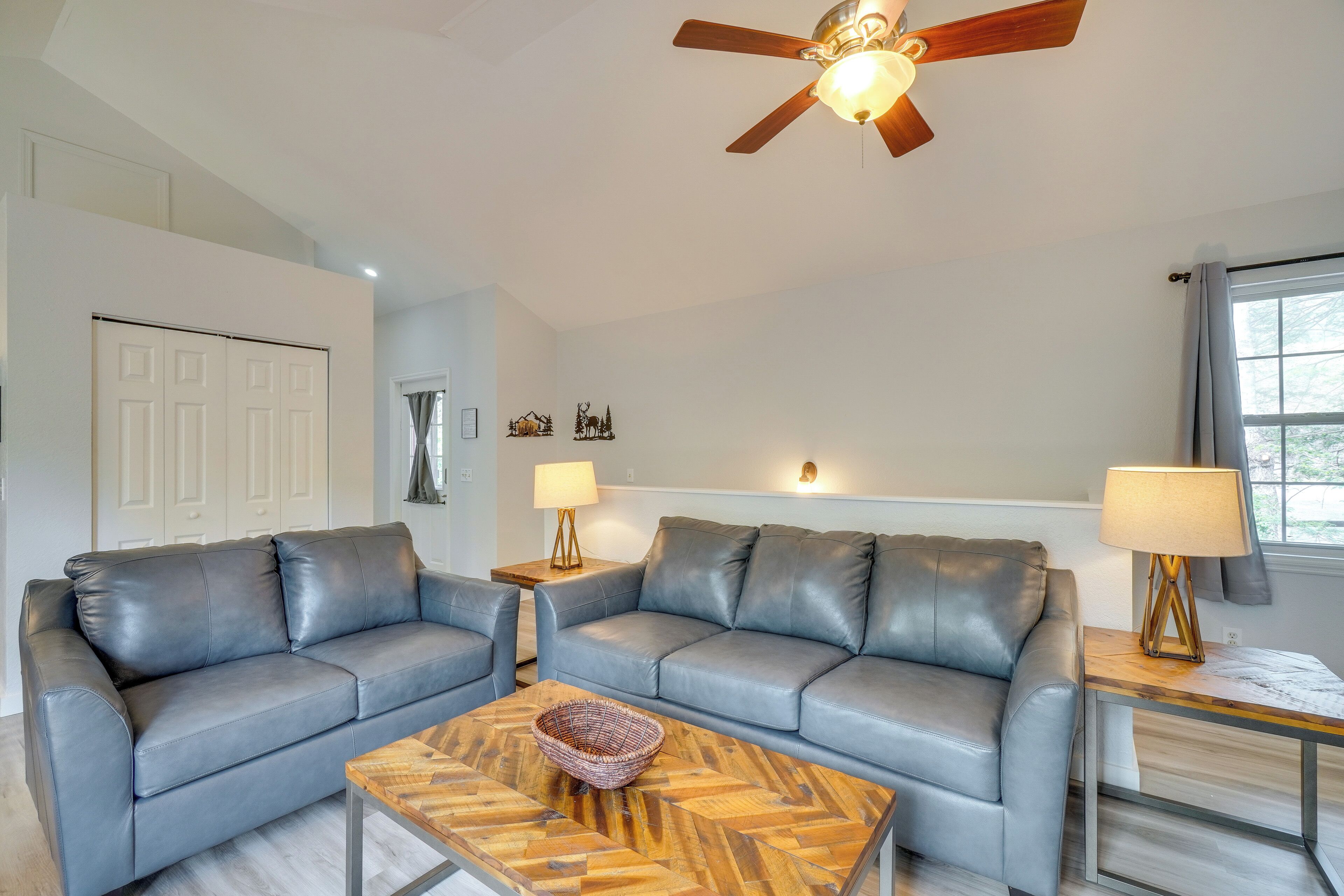 A modern living room with two grey leather sofas, a wooden coffee table, a ceiling fan, and a window with curtains.