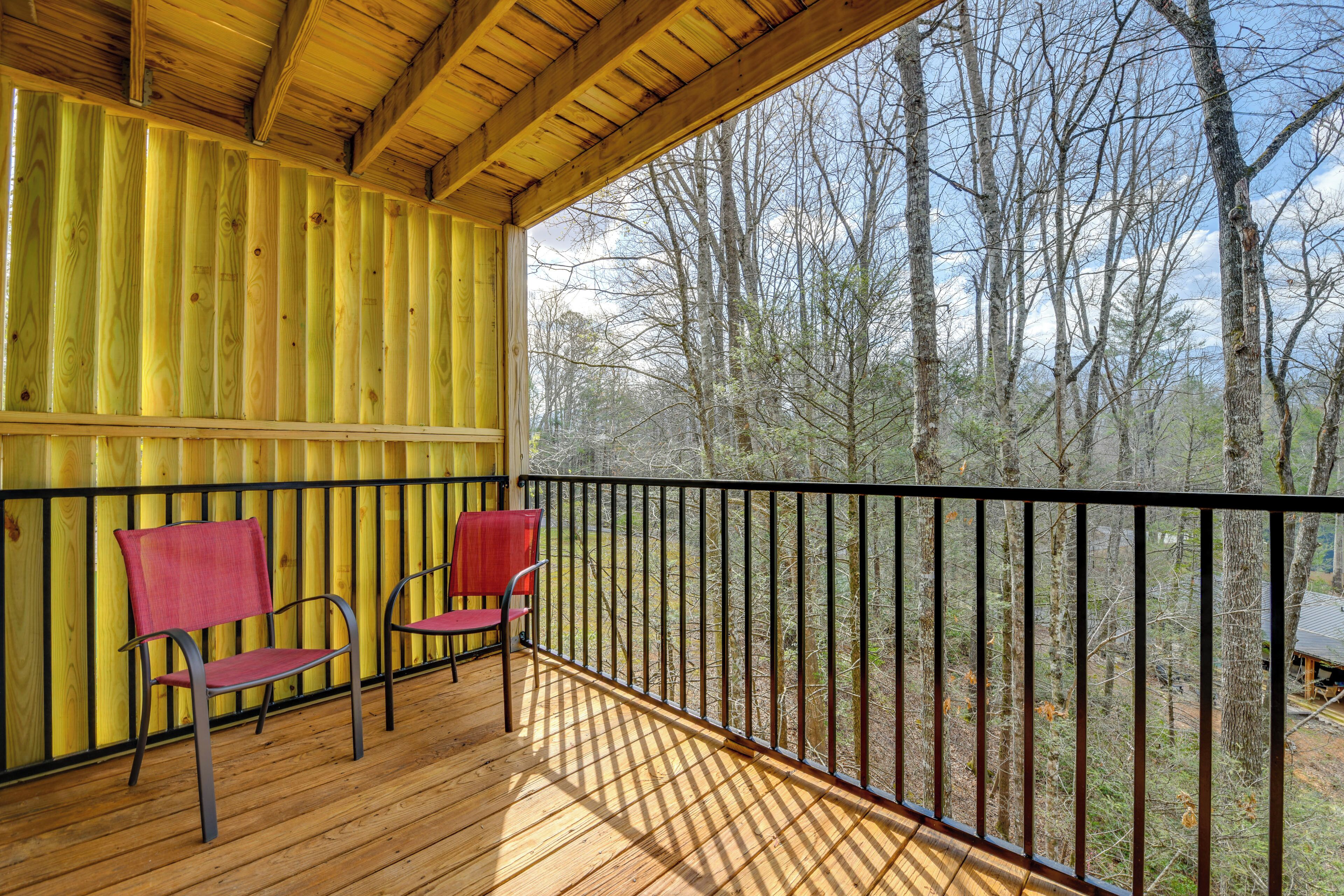 A wooden deck with two red chairs and a black railing overlooking a forest.