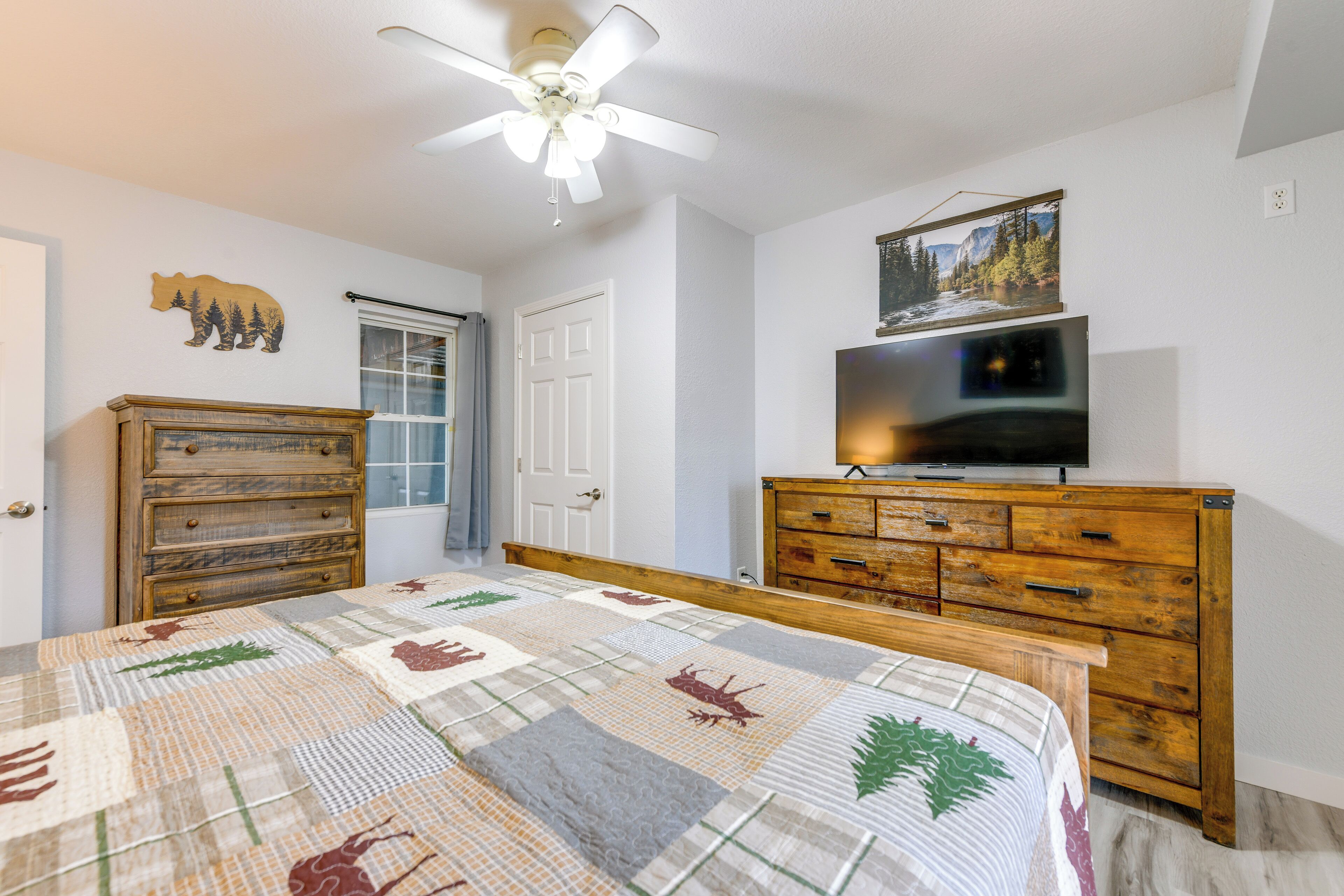 A bedroom with a bed, a wooden dresser, a television, a ceiling fan, a door, and a wall with a bear decoration.