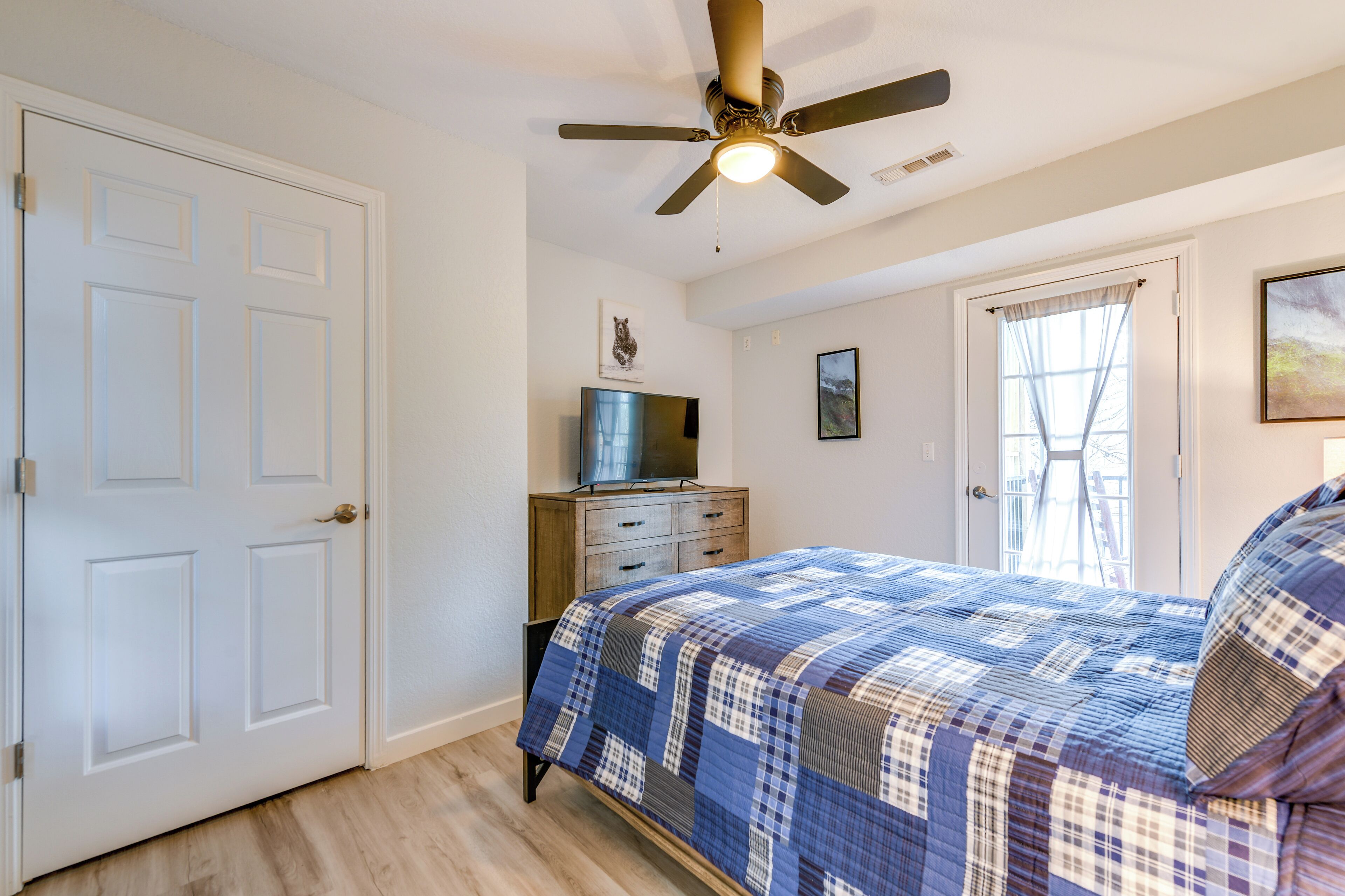 A bedroom with a bed, a ceiling fan, a dresser, a television, and a window with curtains.