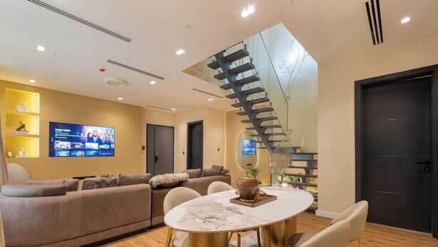 Una sala de estar moderna con un sofá curvo, una mesa redonda con sillas, un televisor montado en la pared y una escalera de caracol que conduce a otro nivel.