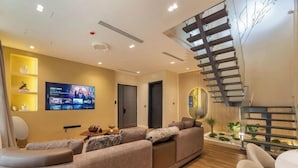 Una sala de estar moderna con un televisor de pantalla plana grande, un conjunto de sofás, una escalera y una pequeña mesa con adornos.