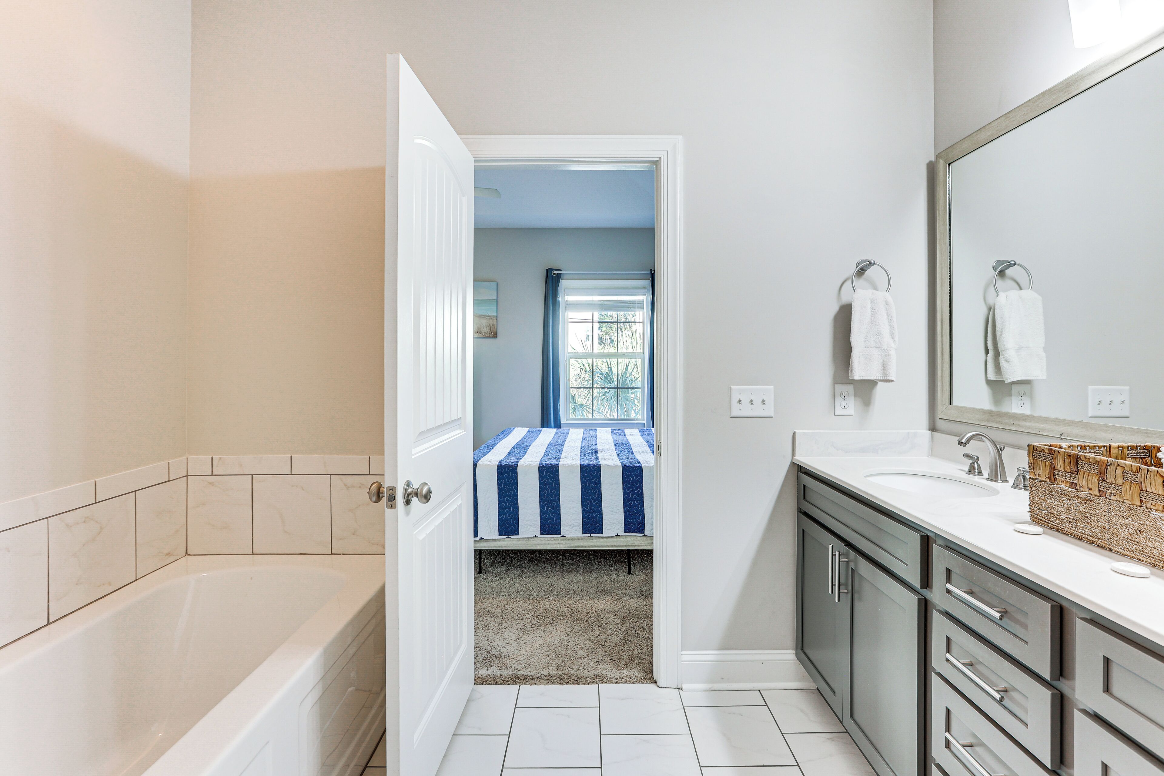Une salle de bain avec une baignoire, un lavabo et un miroir. Une porte donne sur une chambre à coucher qui contient un lit et une fenêtre.