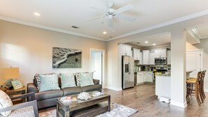 A spacious living room with a sofa, coffee table, and a dining area with chairs. A kitchen with modern appliances is visible in the background. - 3 Mi to Pier Village: Dog-friendly St Simons Home! (St. Simons Island)