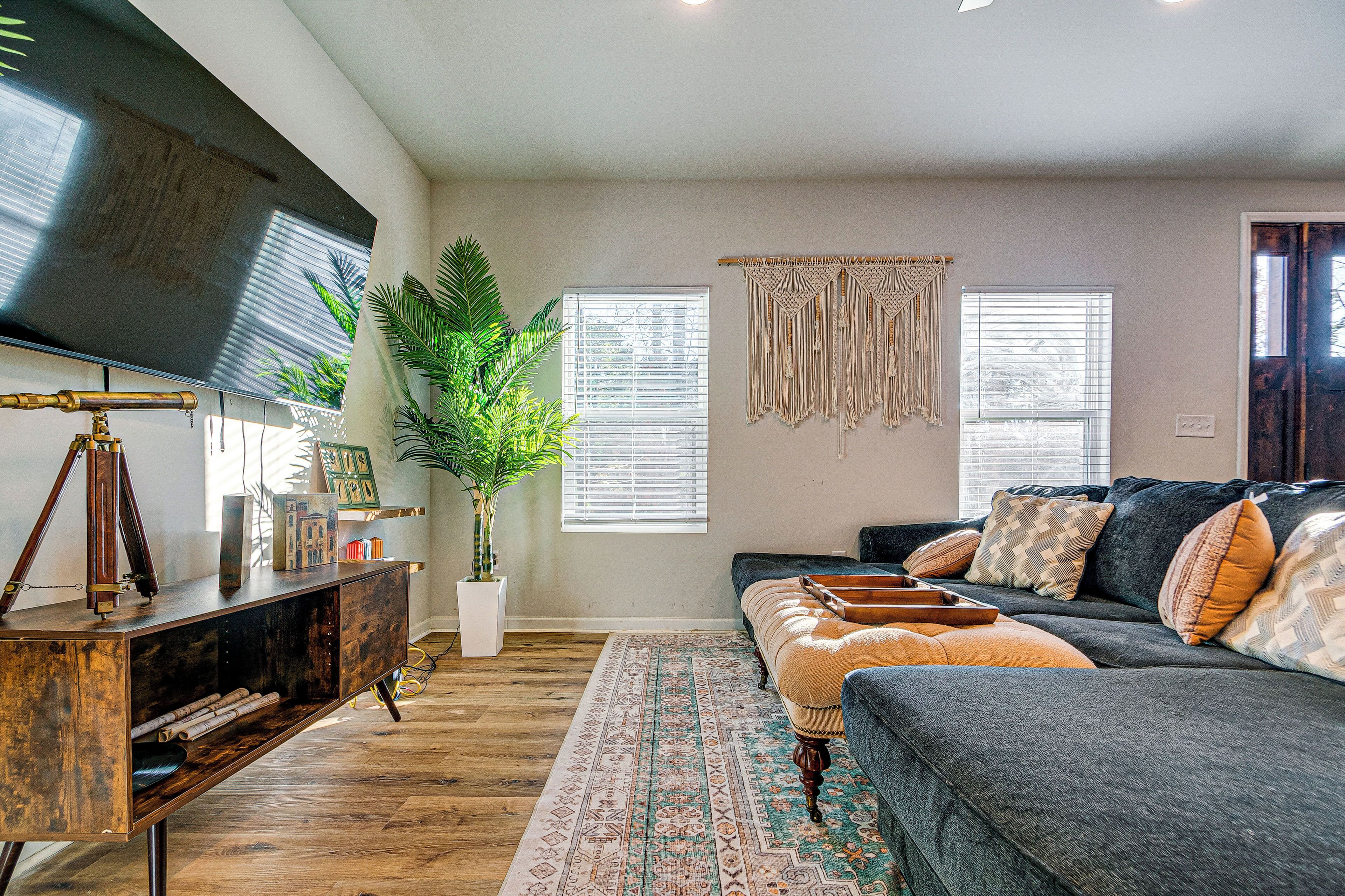 A living room with a dark grey sofa, a wooden coffee table, and a large flat-screen TV mounted on the wall. There is a potted plant and a patterned rug on the floor.