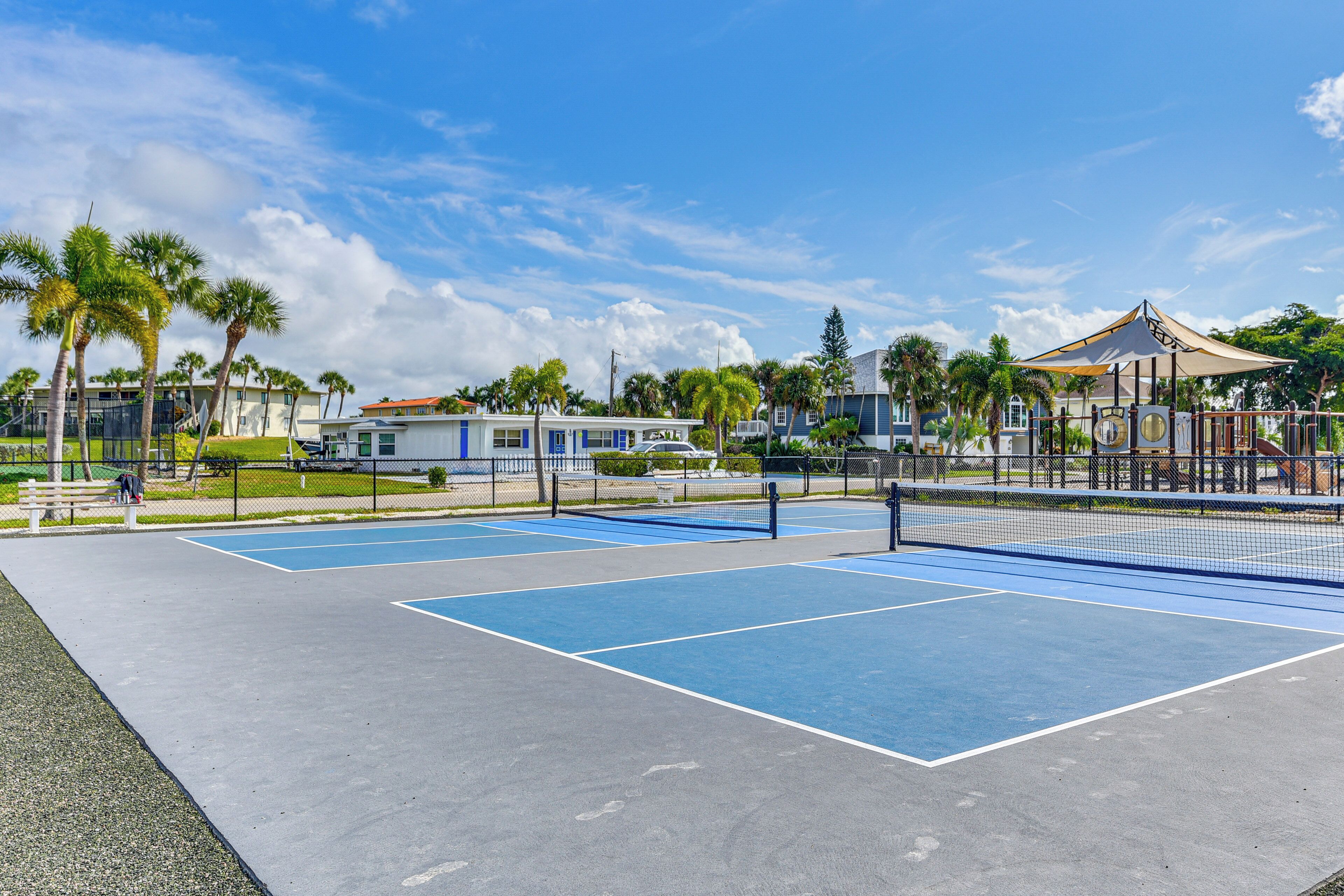 Una cancha de tenis al aire libre con superficies azules y grises, rodeada de palmeras y una valla.
