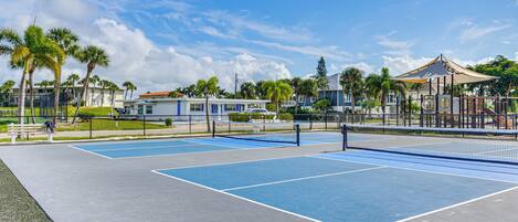 Una cancha de tenis al aire libre con superficies azules y grises, rodeada de palmeras y una valla.