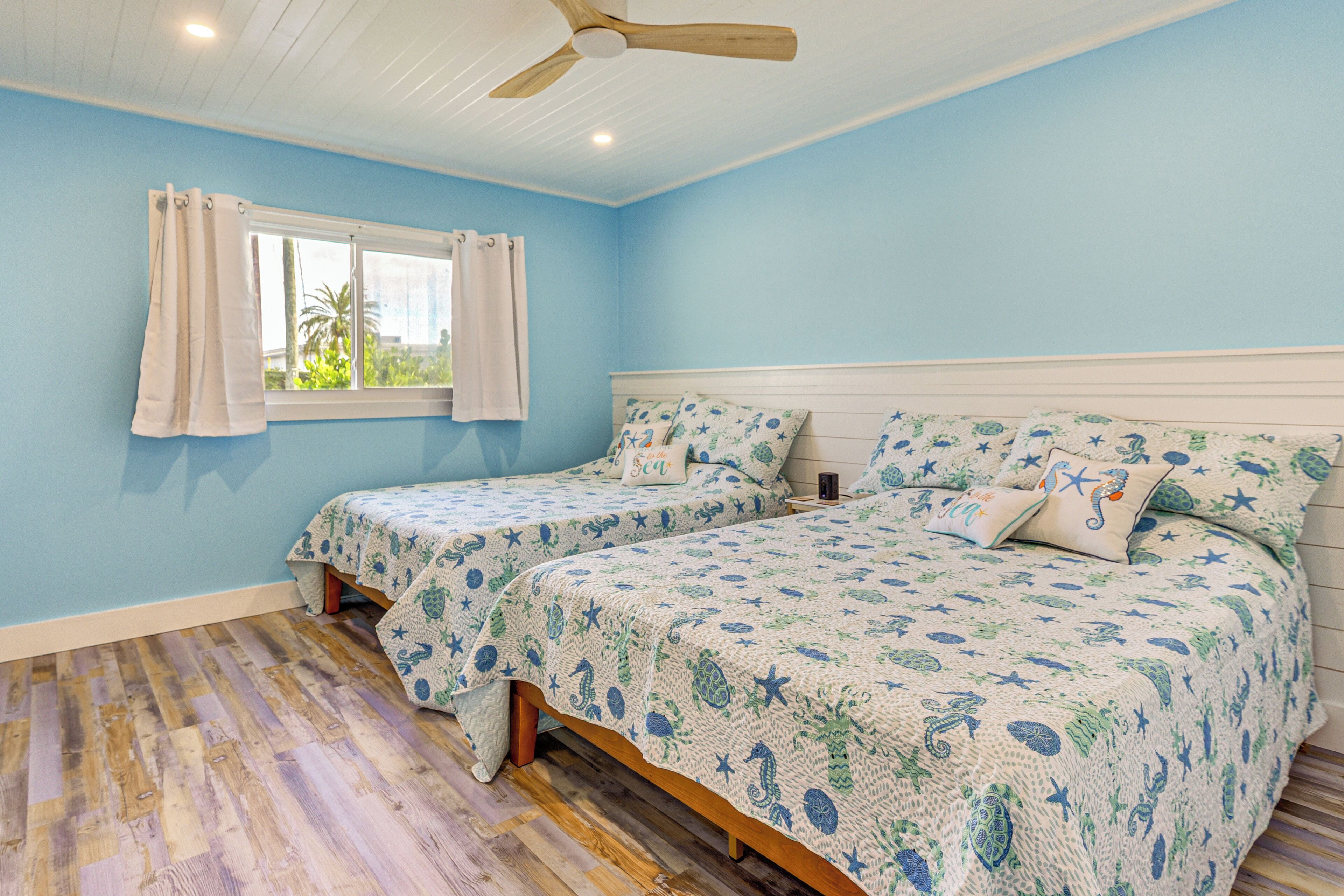Une chambre avec deux lits, une fenêtre avec des rideaux, un ventilateur de plafond et un plancher en bois.