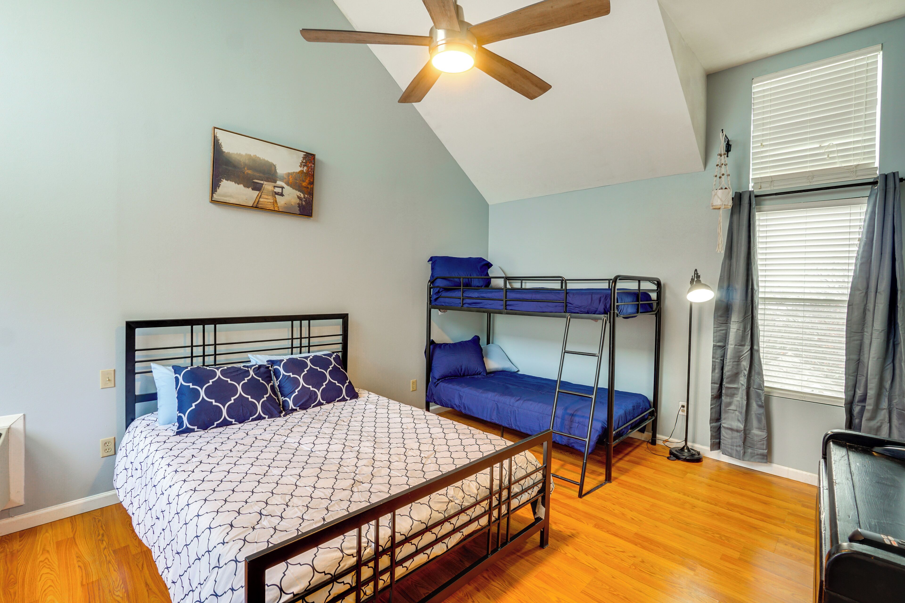 A bedroom with a bunk bed, a single bed, a ceiling fan, a painting, and a window with curtains.