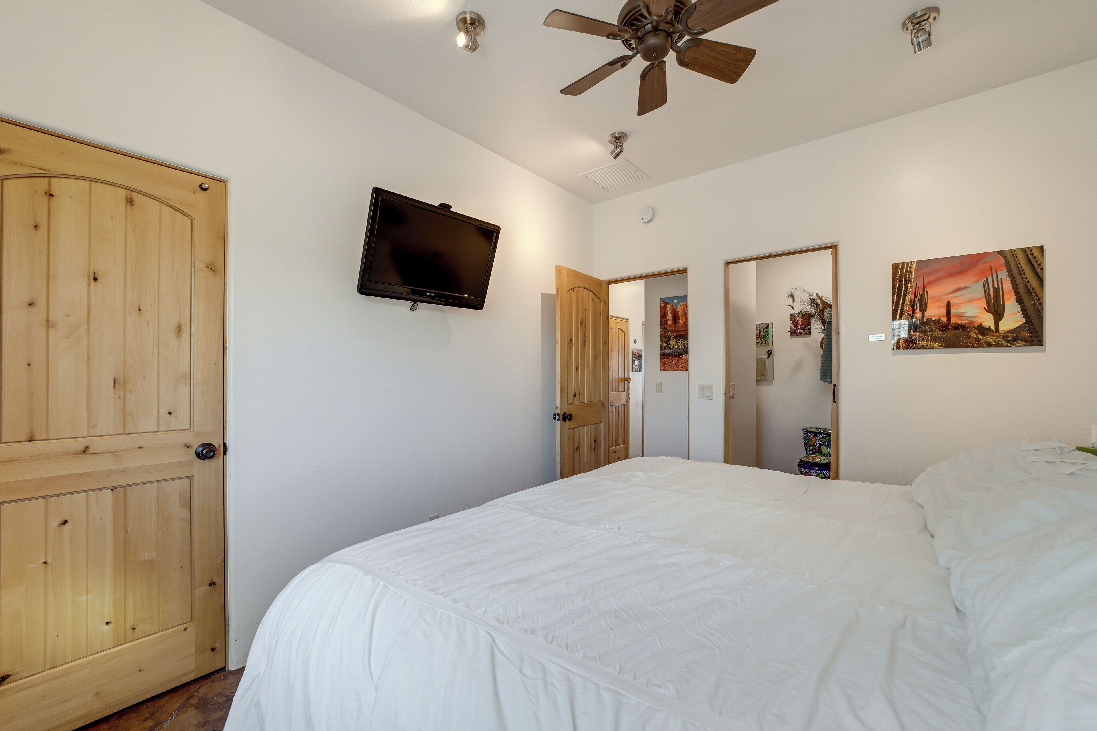 Un dormitorio con una cama grande, puertas de madera, un ventilador de techo, un televisor montado y obras de arte en la pared.