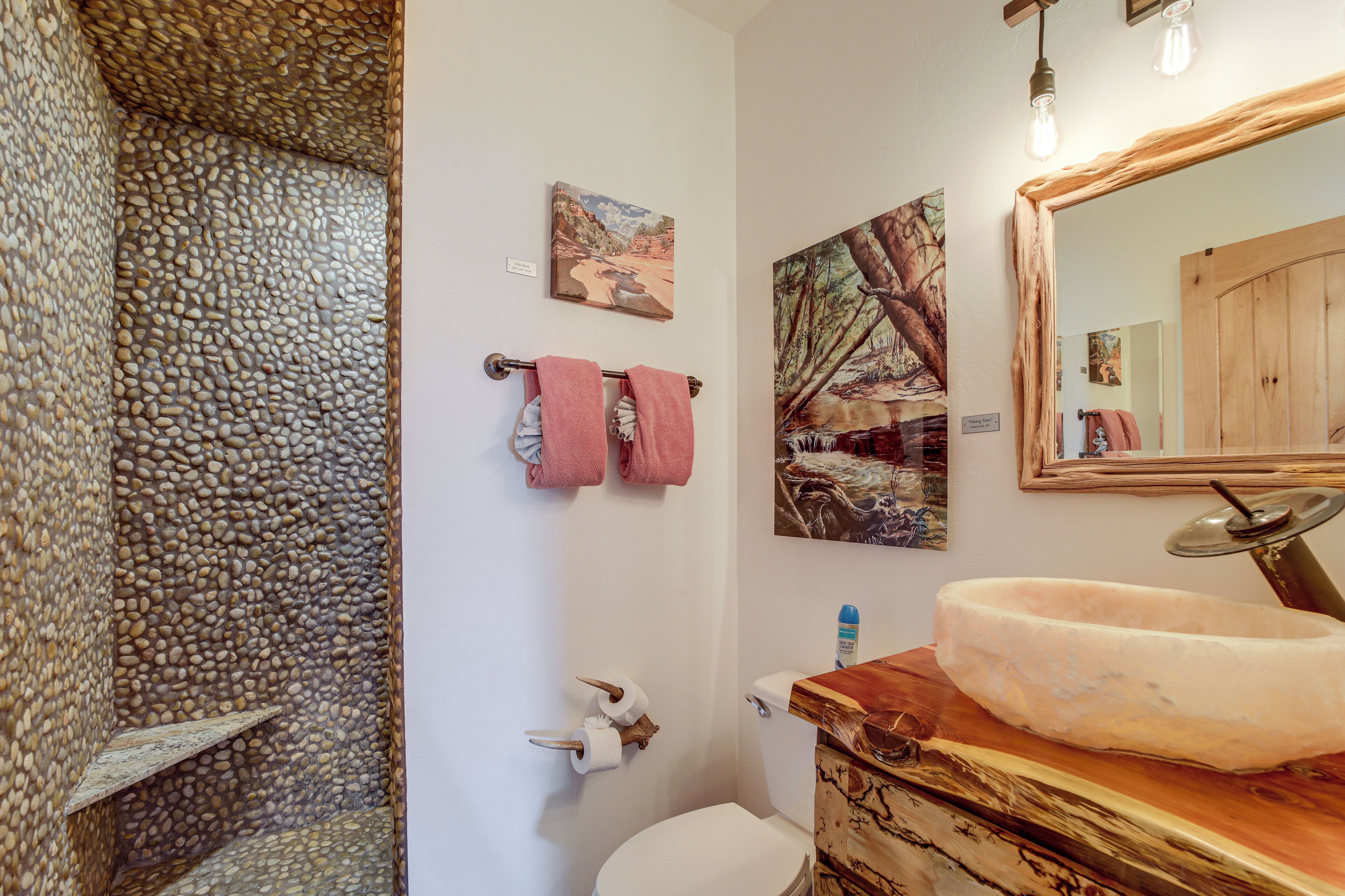A bathroom with a stone wall, a wooden vanity, a white toilet, and two framed paintings.