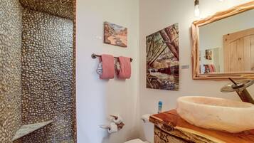 A bathroom with a stone wall, a wooden vanity, a white toilet, and two framed paintings.