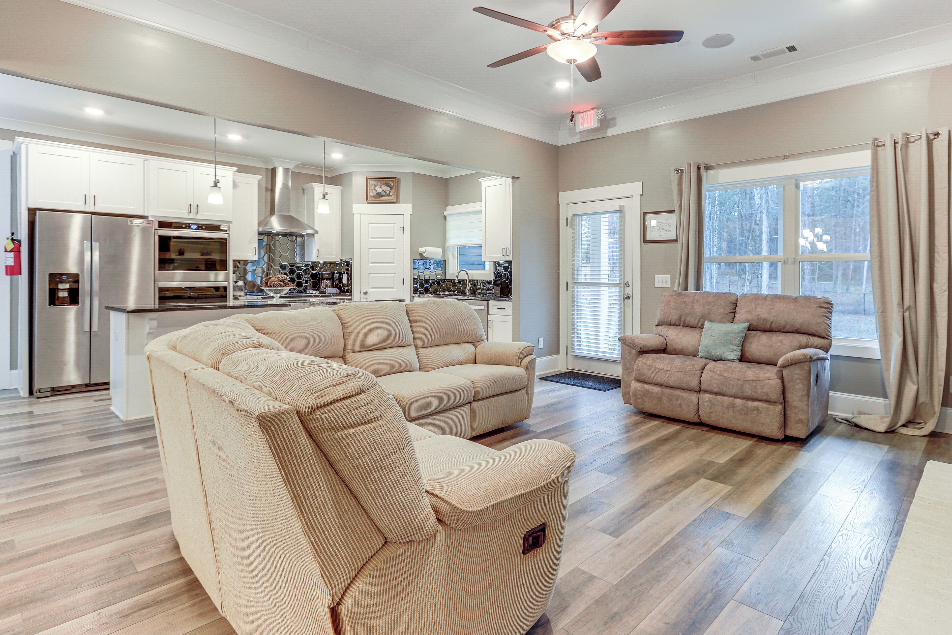 A spacious living room with a beige sectional sofa, a recliner, and a dining area with a kitchen in the background.