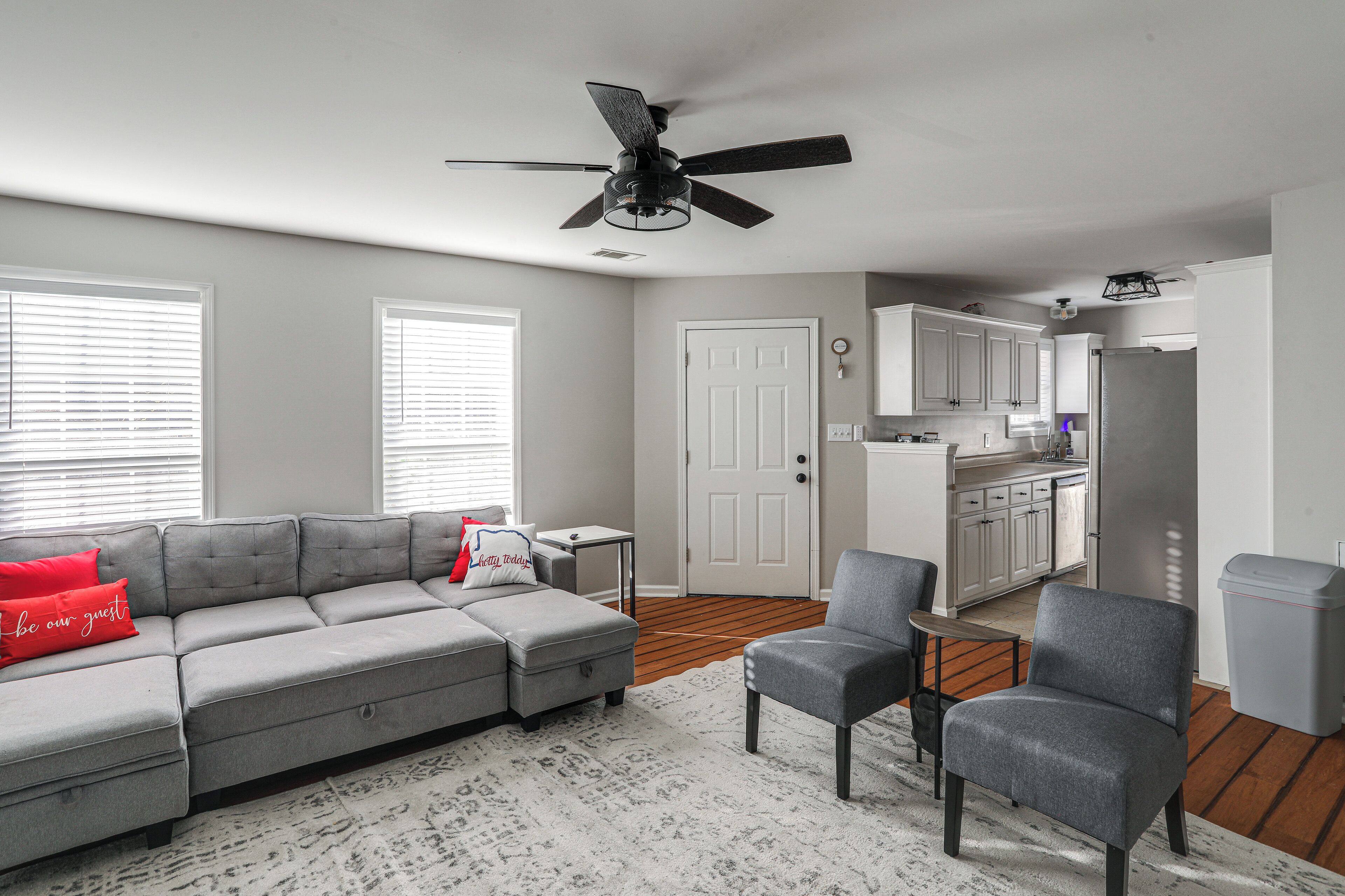 Ein Wohnzimmer mit grauem Ecksofa, einem grauen Sessel und einem weißen Teppich. Es gibt einen Deckenventilator, eine weiße Tür und eine Küchenzeile mit weißen Schränken und einem Kühlschrank.