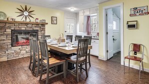 Un coin repas avec une cheminée en pierre, des chaises en bois, une table dressée pour quatre personnes et, au fond, une cuisine.