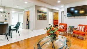 A spacious living area with a glass coffee table, a flat-screen TV mounted on the wall, and a dining area with a table and chairs. - Sand Volleyball Court, Pool, Hot Tub! Fun Retreat (Port Wentworth)