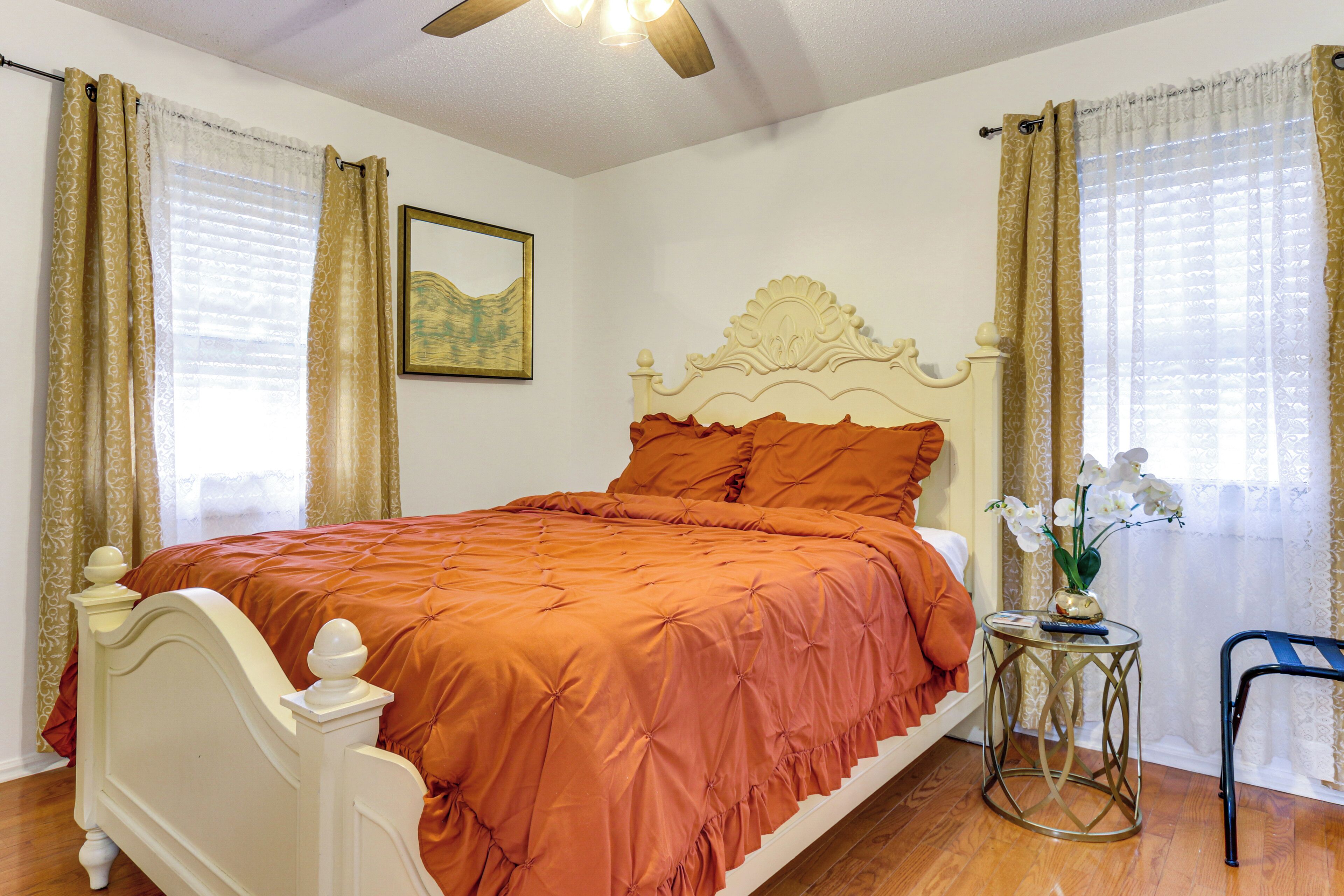 A bedroom with a bed, curtains, a framed picture, a small table with a vase, and a chair.