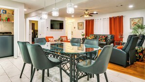 A modern living room with a glass-top dining table surrounded by chairs, a sofa, and a recliner. There is a ceiling fan, a TV mounted on the wall, and a window with curtains. - Sand Volleyball Court, Pool, Hot Tub! Fun Retreat (Port Wentworth)