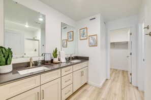 Un baño moderno con un espejo grande, dos lavamanos y una ducha. Hay cuadros enmarcados en la pared y una planta en maceta sobre el mostrador.