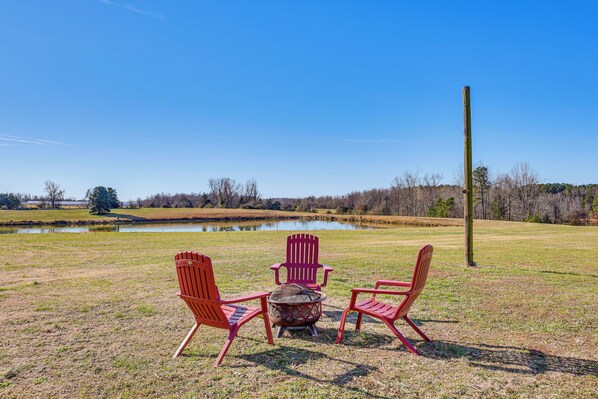 A grassy field with a pond, three red Adirondack chairs, and a small fire pit.