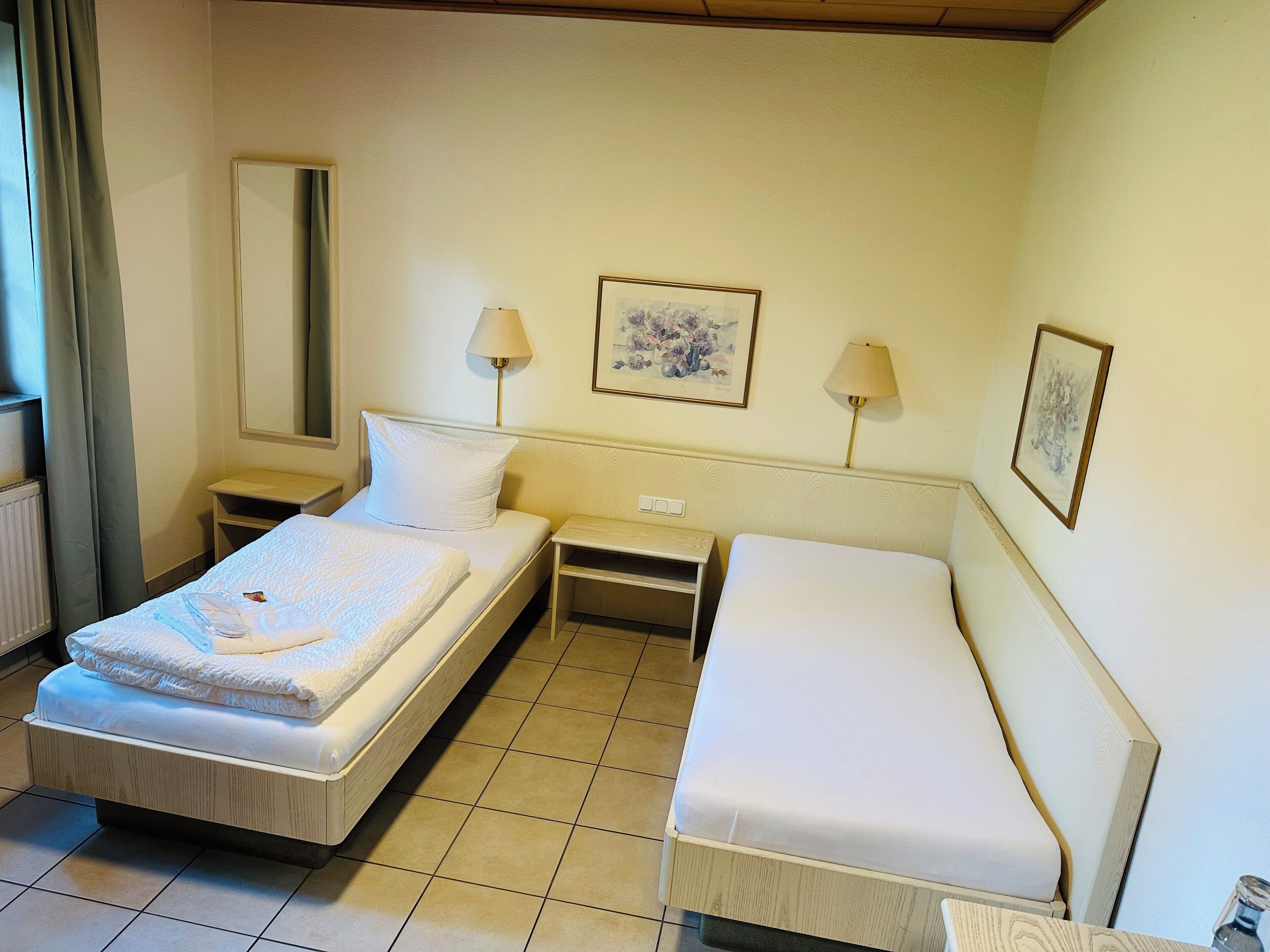 Une chambre d’hôtel avec un lit simple, un petit lit, deux tables de chevet, une commode, une lampe, un miroir et deux tableaux encadrés accrochés au mur.