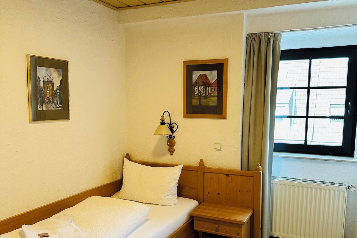 A single bed with a wooden headboard, a bedside table, and a nightstand. There are two framed pictures on the wall, a window with curtains, and a radiator.